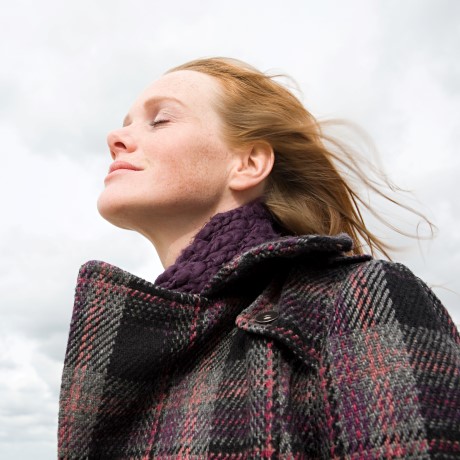Woman with eyes closed, standing outside with a coat on, with wind blowing in her hair.  