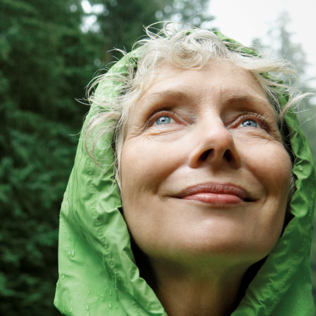 Woman wearing a green rain jacket, standing in a forest and looking up at the sky.