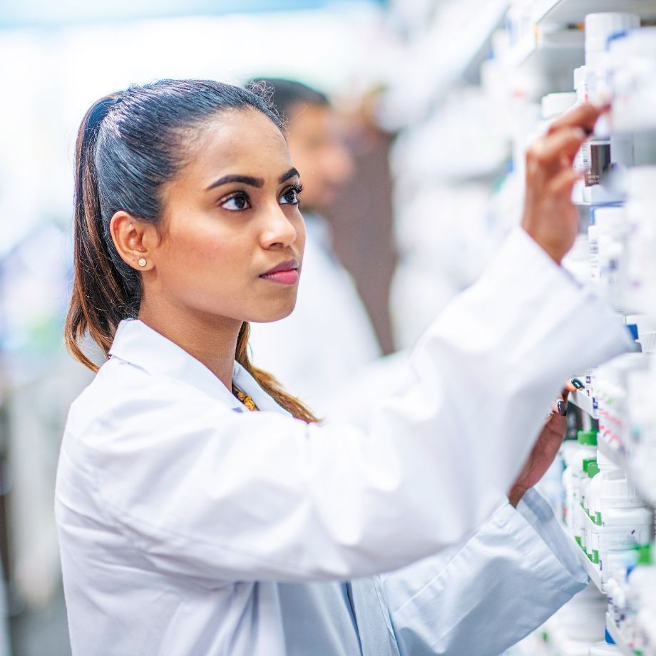 Woman in white lab coat reaching for a bottle of pills on a shelf